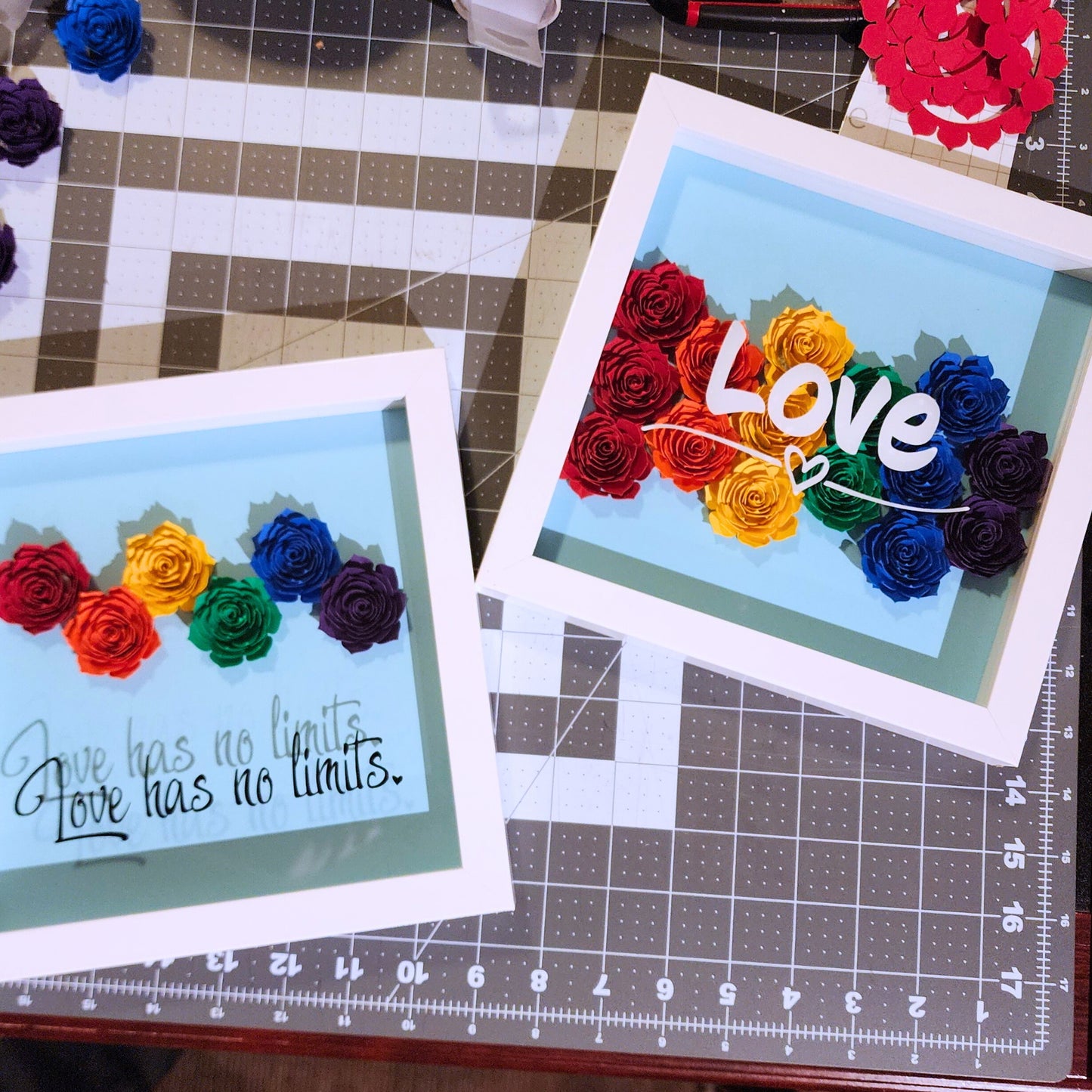 Two framed shadow box artwork with rainbow-colored paper roses. One says 'Love has no limits' black vinyl text on a blue background. The Other says 'Love' in white vinyl text on a blue background.