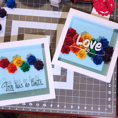 Two framed shadow box artwork with rainbow-colored paper roses. One says 'Love has no limits' black vinyl text on a blue background. The Other says 'Love' in white vinyl text on a blue background.